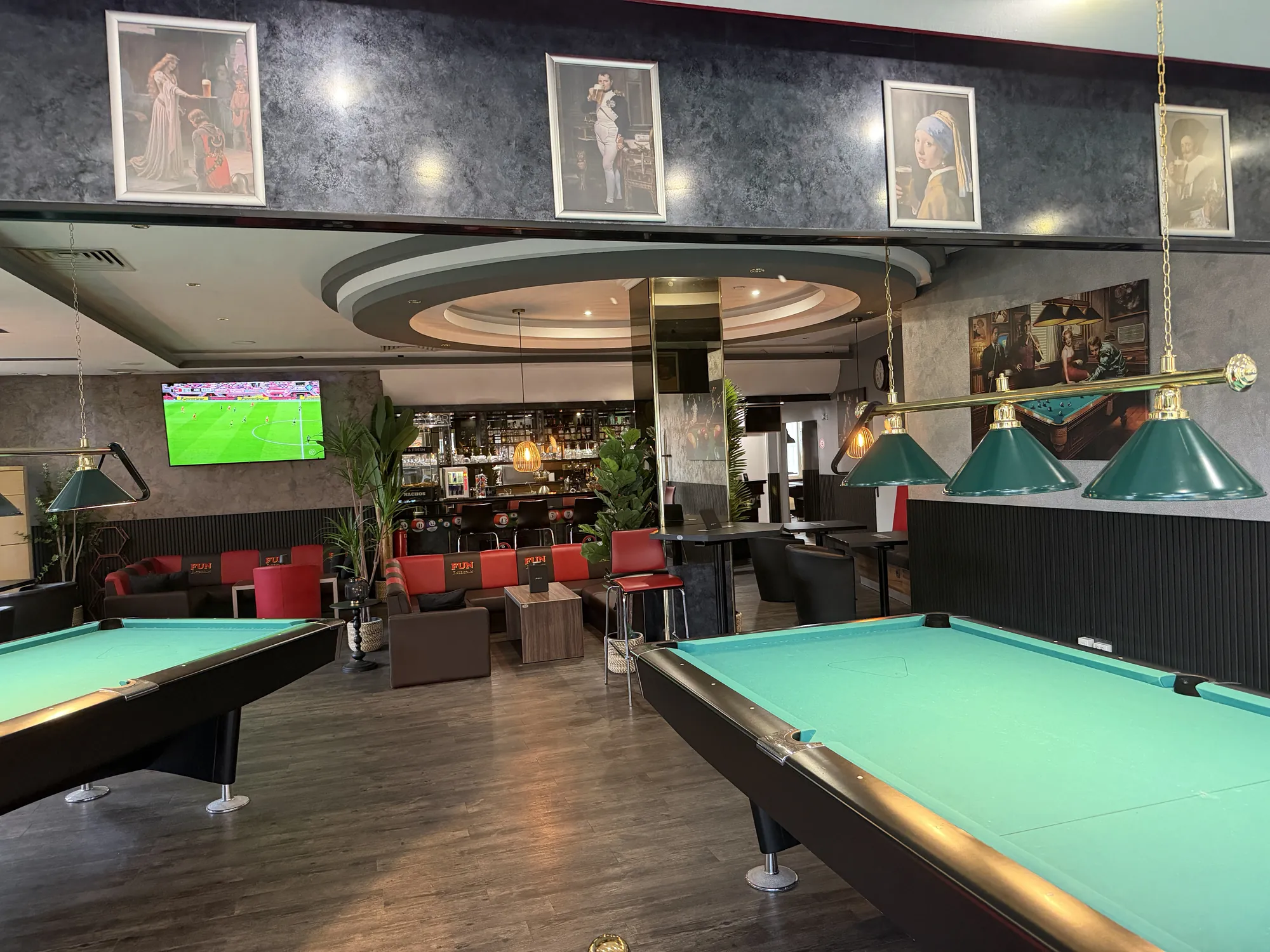 Zwei Billardtische mit grünem Tuch unter Messinglampen in der Players Lounge Dorsten, dahinter Lounge mit roten Sofas und die Bar